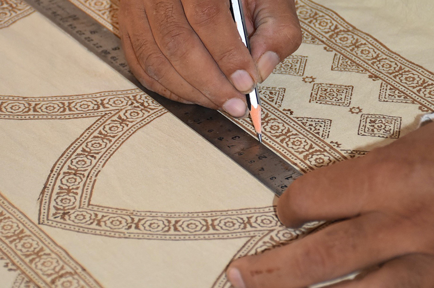 A man is using a pencil and ruler to draw an Indian pattern on a large piece of beige fabric.