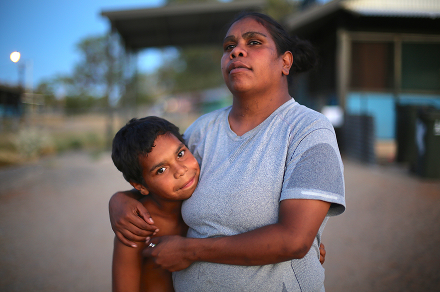 Still from the film In My Blood It Runs. A woman and a young boy stand with their arms around each other. The boy is giving the camera a small smile, while the woman looks off into the distance.