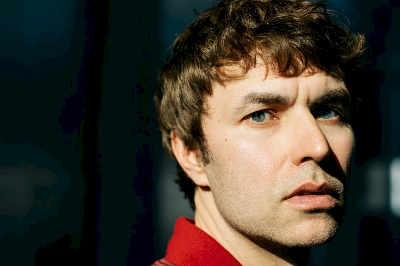 A photo of Karol Radziszewski. Only his face is in the photo. He is stood against a black background, looking off camera to the left.
