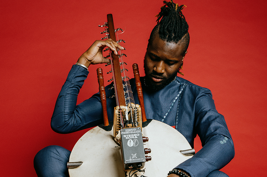 Suntou Susso sat down against a red background holding a Kora.