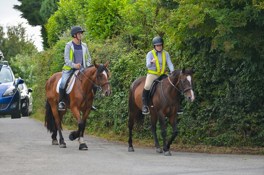 Young couple ride their horses along road wearing safety gear and being seen by other road users , a queue of cars are waiting behind needing to pass when it is safe to do so.