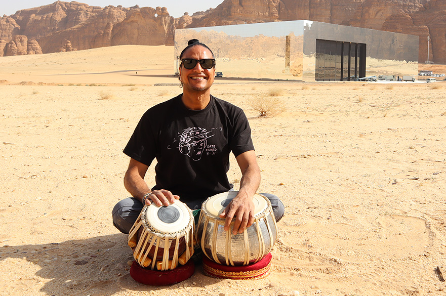 A male musician sat on the floor in the desert with two tabla drums.