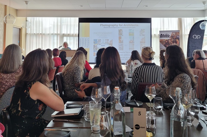 People at a networking event sat at tables watching someone present in front of a screen.