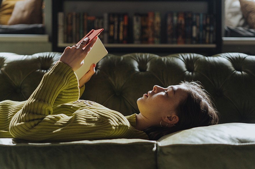 A women laid on a sofa reading a book.