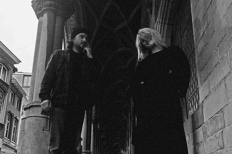 A black and white image of a man and a woman stood outside under an arched roof.