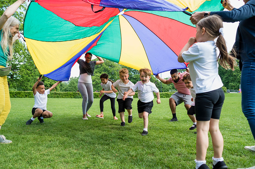 Children in school PE kit with teachers playing with a parachute outside