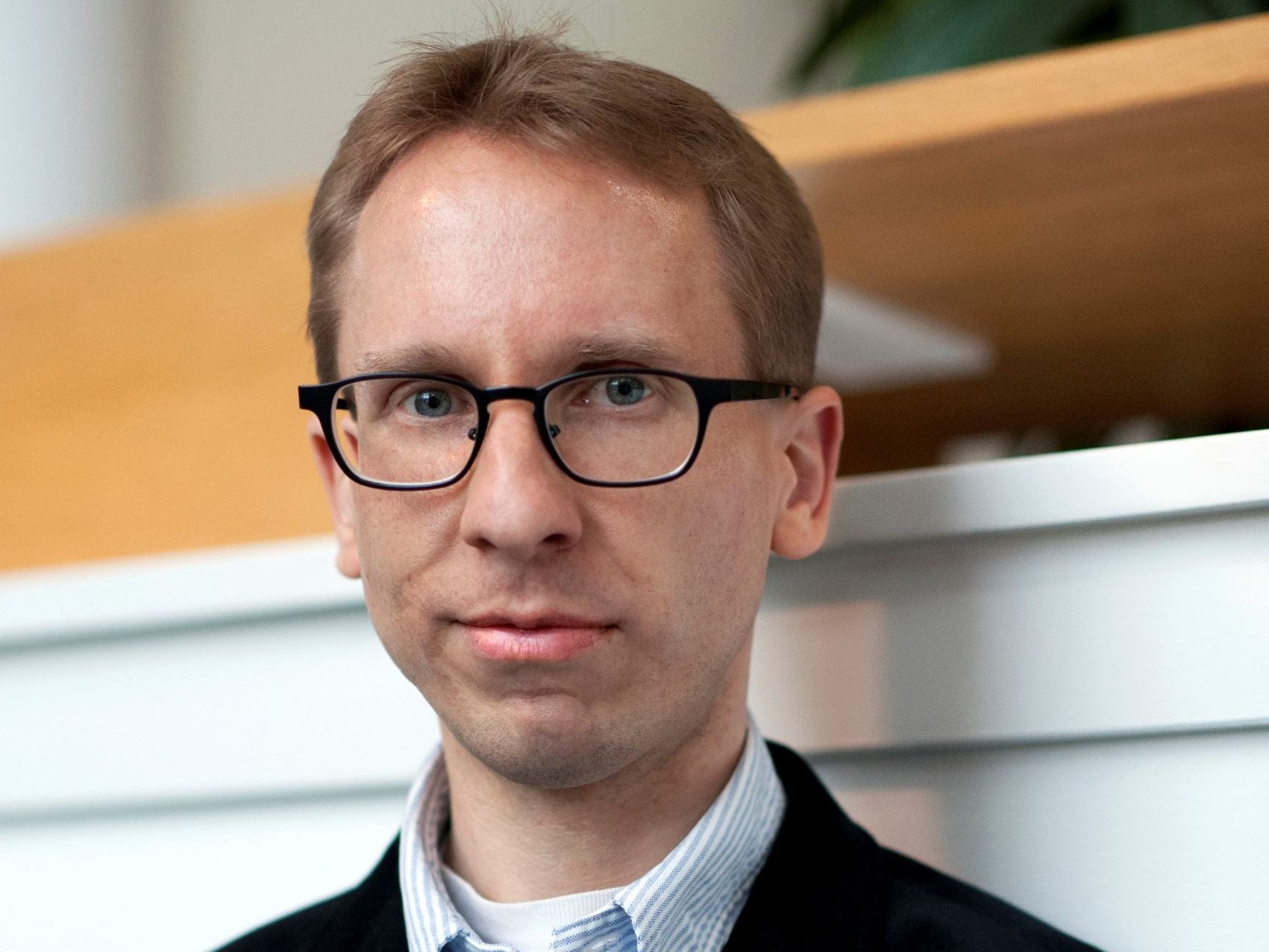 Person wearing glasses, a dark blazer, and a light blue striped shirt. The background is neutral and uncluttered