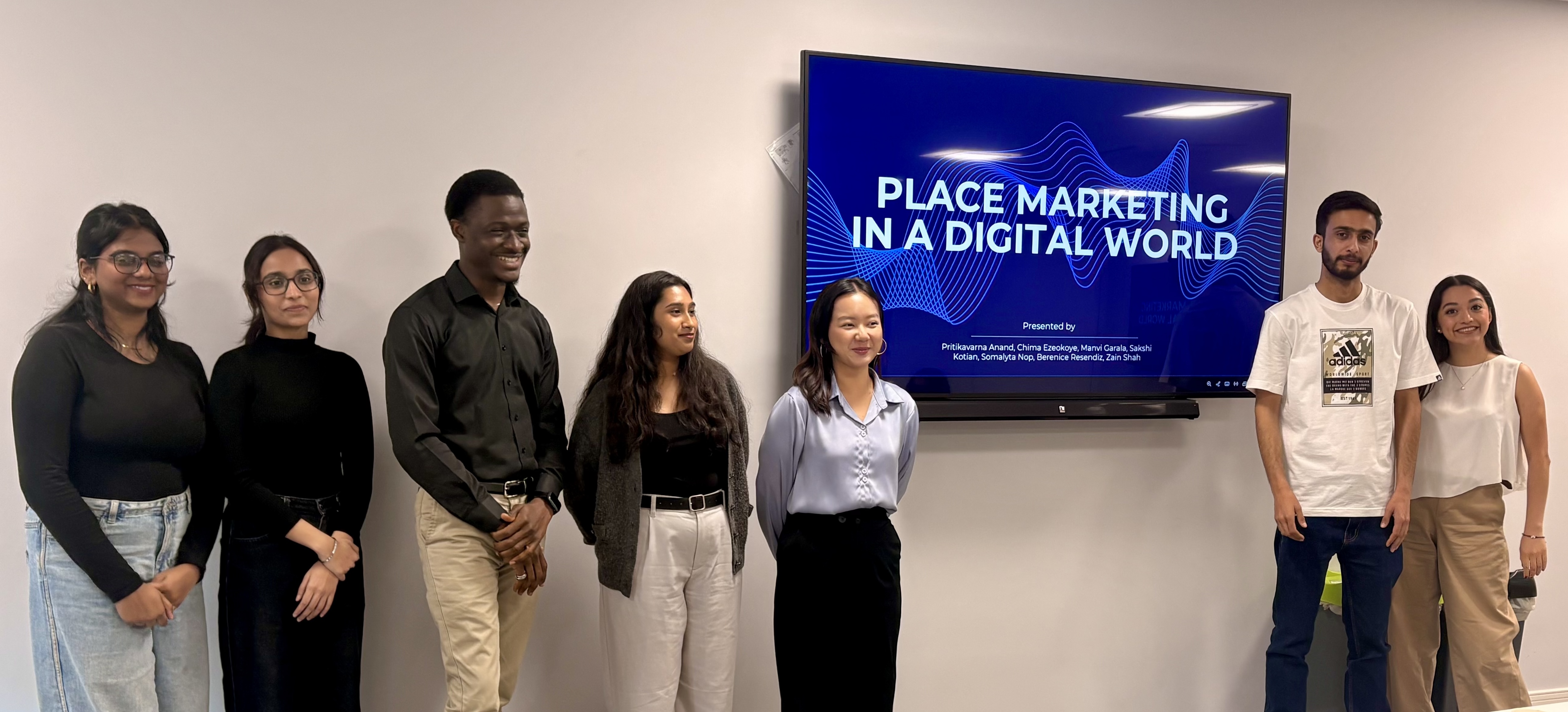 Seven students standing in a classroom in front of a TV screen. 