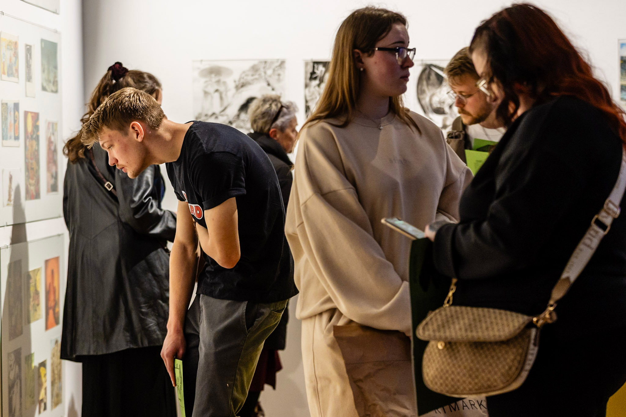 A group of people in an art gallery observing and discussing framed artworks on the walls, with some individuals holding brochures or booklets.