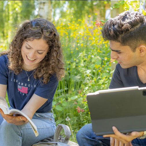 Two students sat on a bench at Brackenhurst campus. One shows the other a page in a book, while the other holds a tablet.