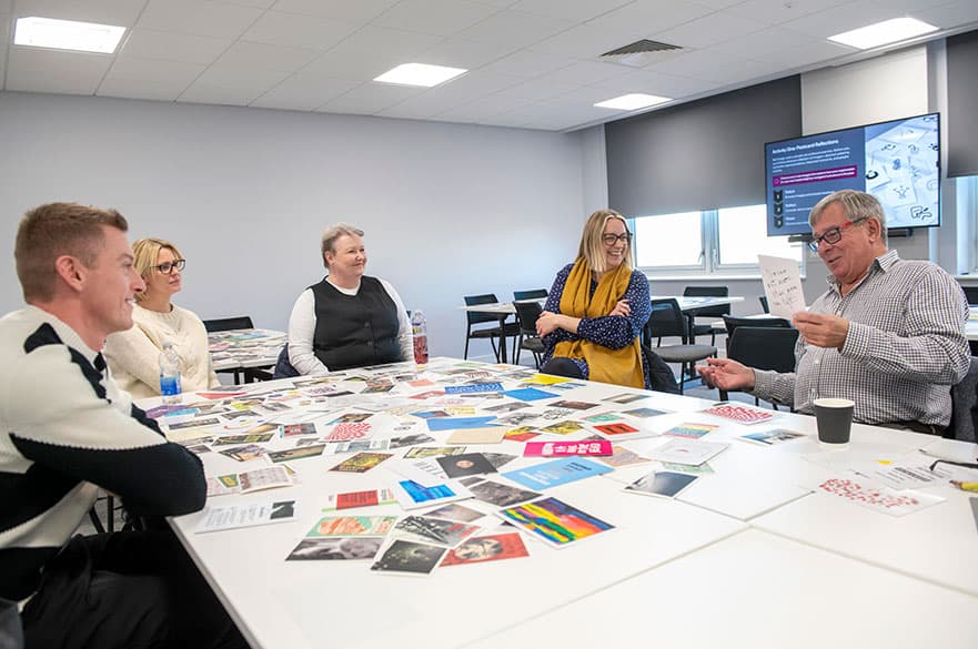 A group of MBA students having a discussion around a table with lots of colourful materials scattered on the table.
