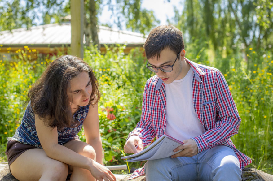 Two students discussing notes in a notebook while sat on a bench outside in the sun at Brackenhurst Campus.