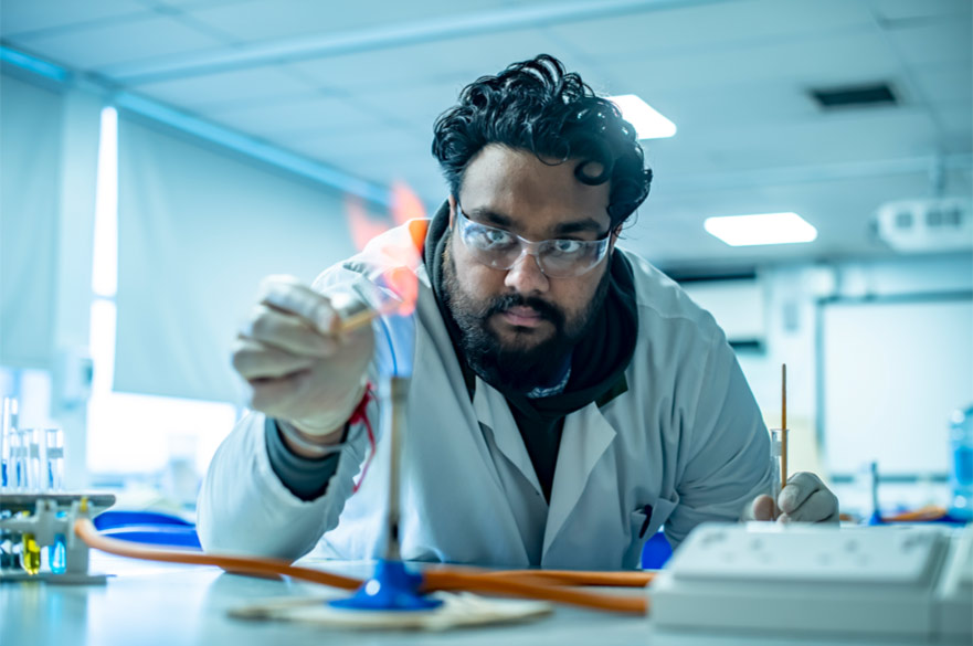 A scientist wearing lab goggles and a lab coat concentrating as he holds an object over a bunsen burner.