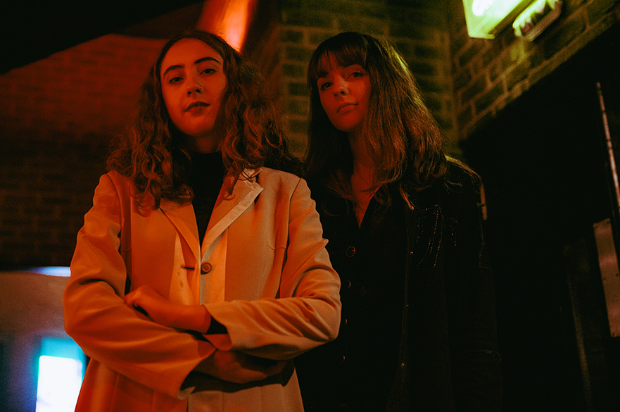 The members of Let’s Eat Grandma stood outside in jackets at night..