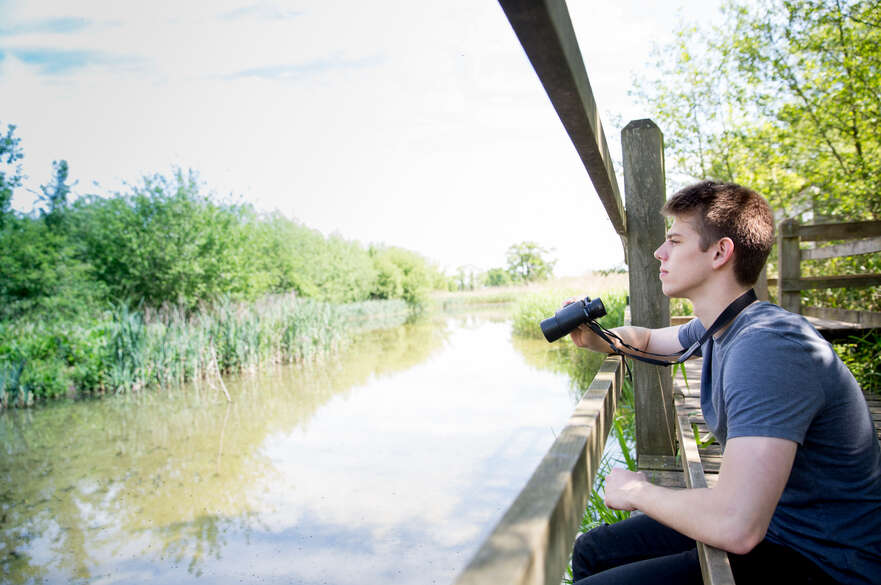 A student looking out over a river, holding a pair of binoculars.