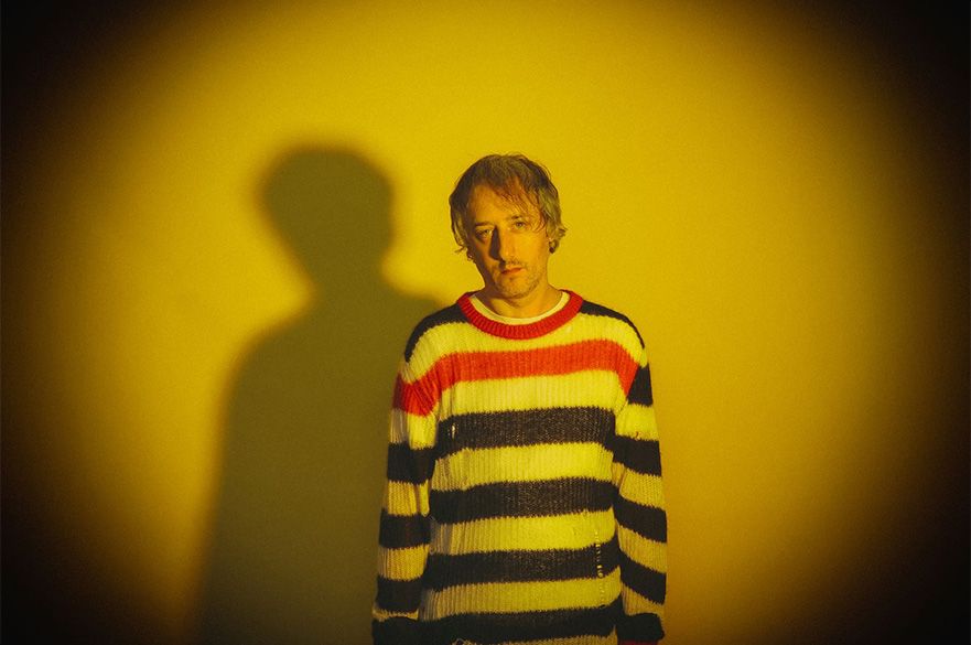 A man wearing a stripy jumper stood looking at the camera with a yellow tinted light shining on him.