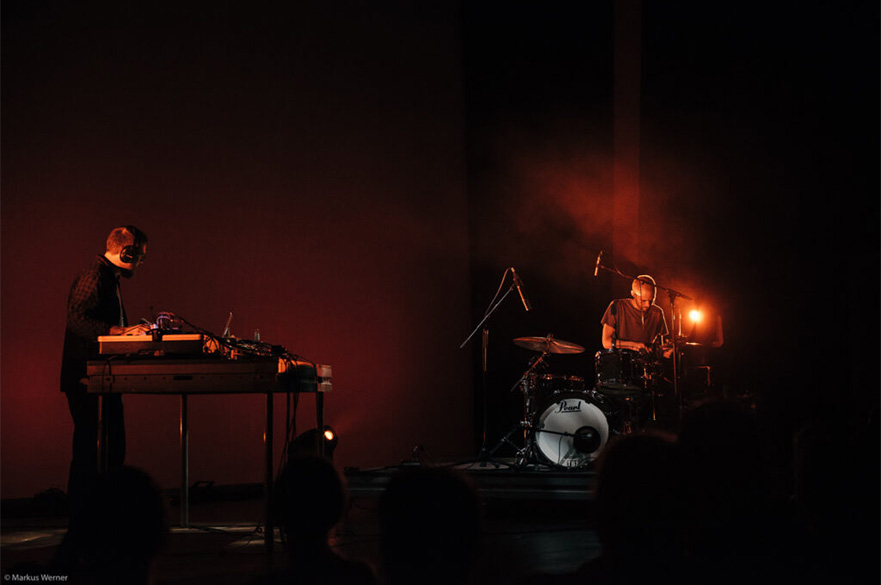 A dark red coloured image of a drummer and DJ on stage.