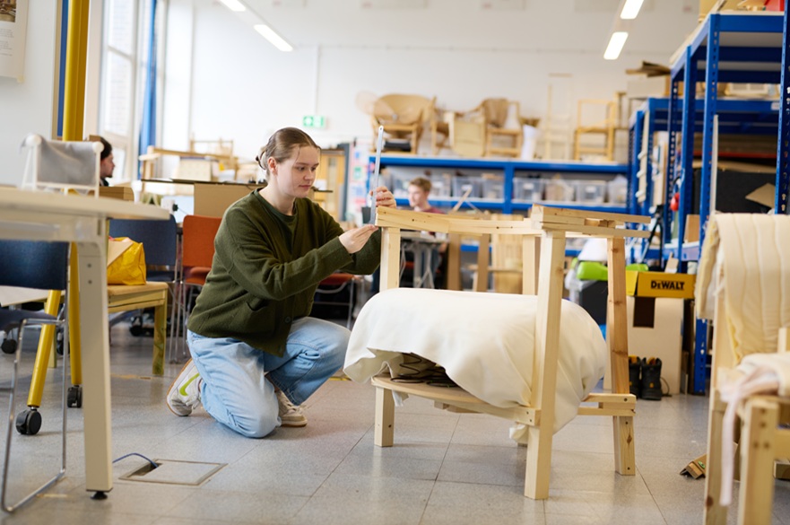 A student at work measuring a wooden chair she has designed. She is kneeling by the chair. She is in a studio.