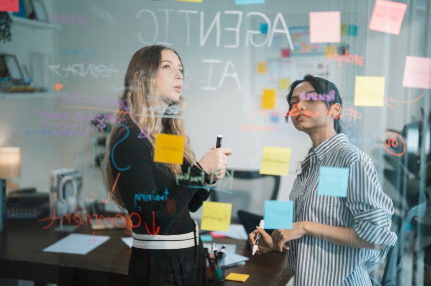 Two young women looking at post it notes and drawings about AI on a board