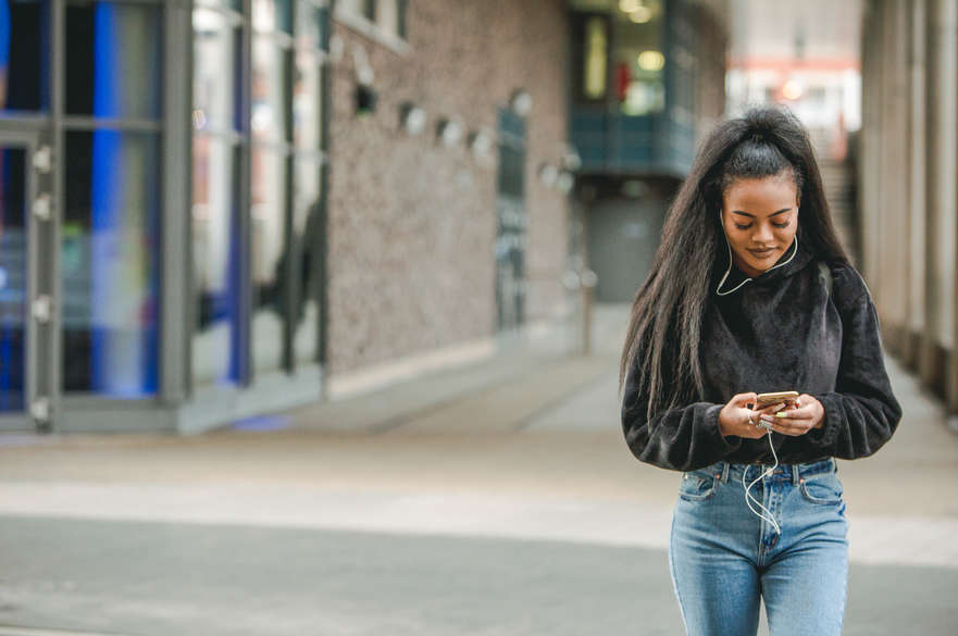 A girl walking along with headphones in, looking at her phone on the City campus