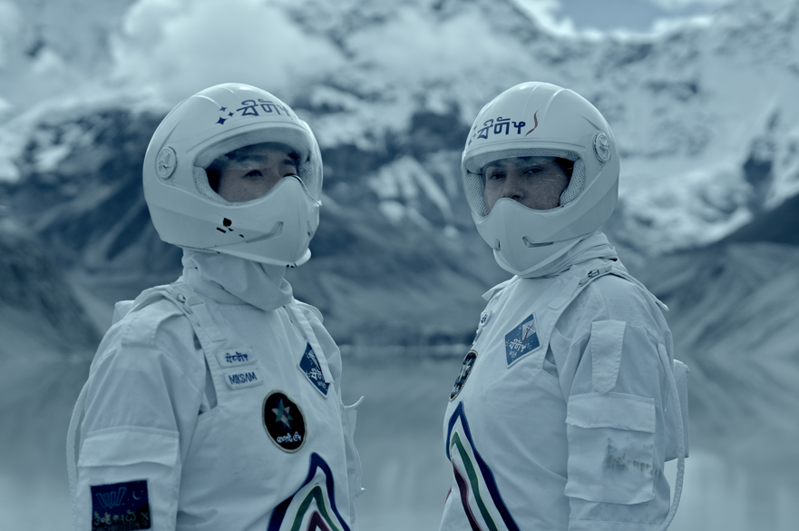 Two people in spacesuits standing on a snowy mountainous landscape