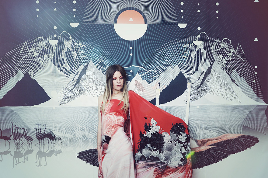 A woman stood in front of a mountain landscape background in a red patterned dress and cape.