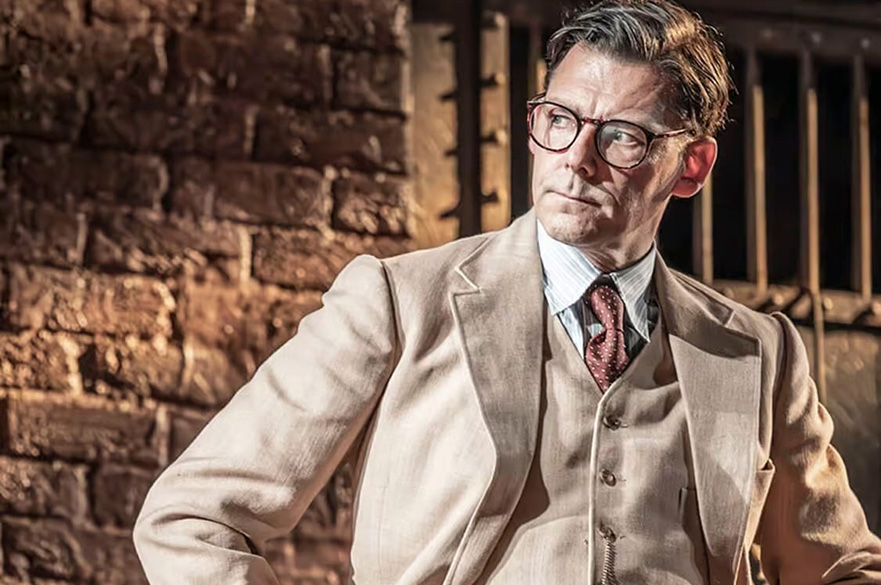 A man in a beige suit, glasses, and a red tie sits in front of a brick wall and metal bars, looking to the side.