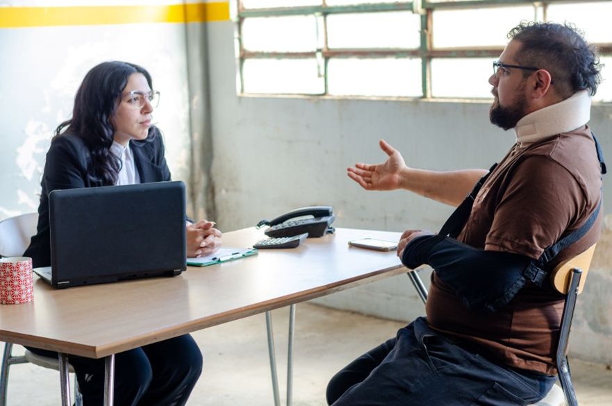 A female lawyer sits across the desk from a male client who is talking to her, his arm is wrapped in a sling