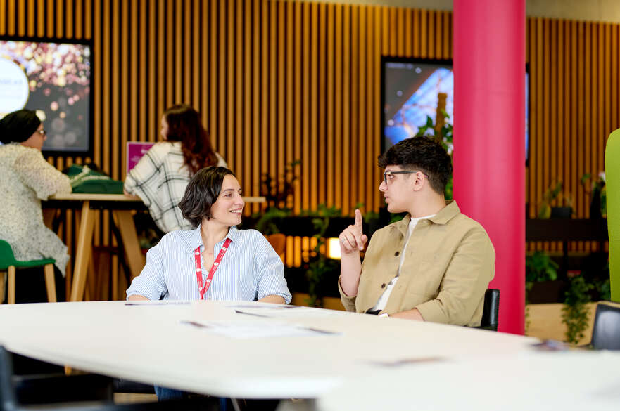 Two people are engaged in a discussion while sitting at a table in a modern office environment with colleagues working in the background.