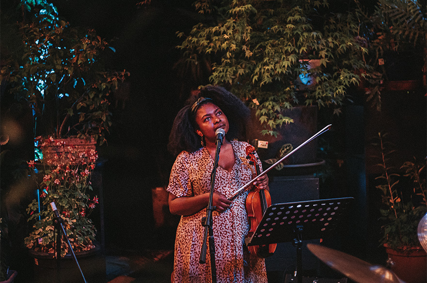 A woman holding a violin with an array of plants and trees behind her.