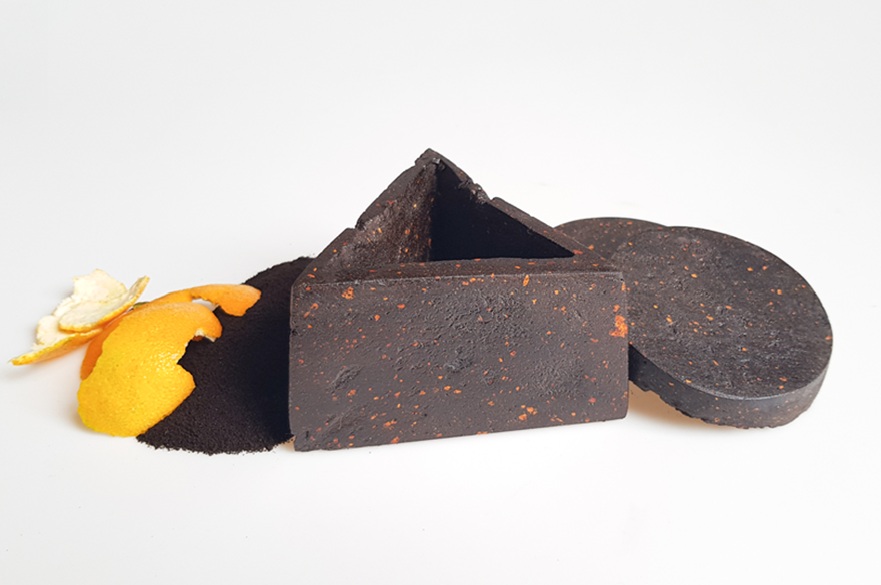 A container made out of recycled coffee grounds next to a disc of the same and some orange peel.