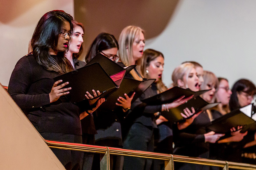 Women singing in a choir on a balcony holding lyric sheets.