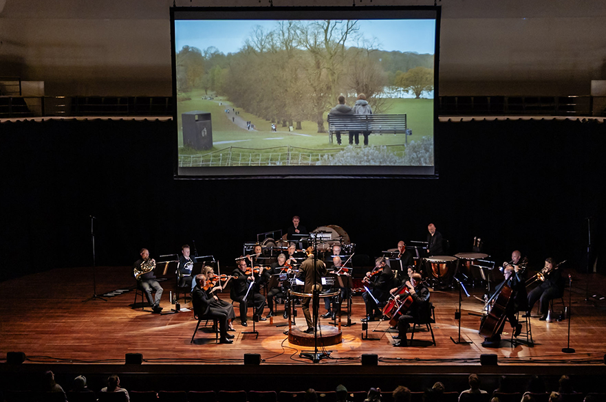 A small orchestra playing on stage in front of an audience, with a large screen suspended above them showing a film - the frame is two people sat on a bench at Wollaton Park.