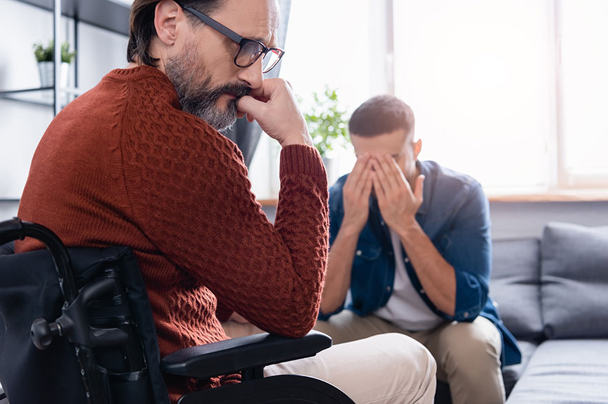 upset disabled man near depressed son obscuring face with hands on blurred background