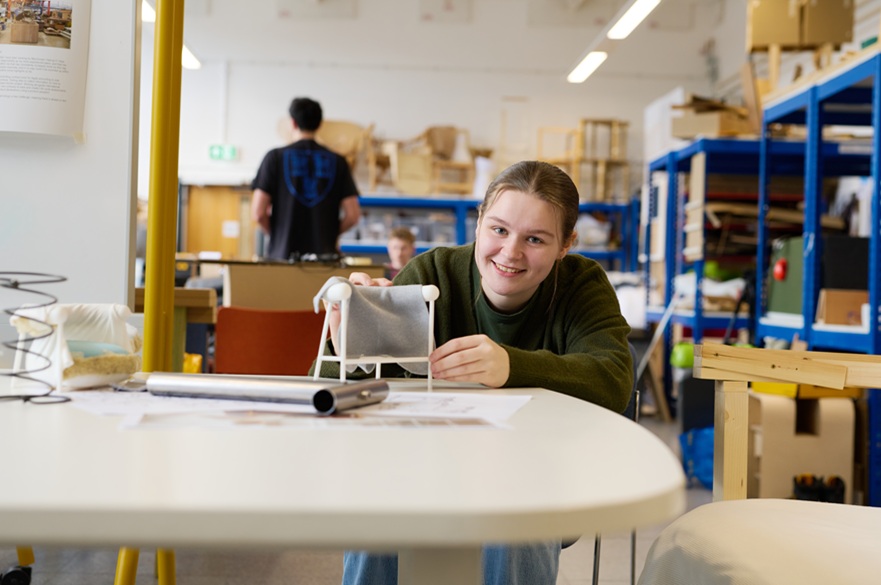 A person looking at the camera to have their picture taken. They are at a work table with a model of their chair design.