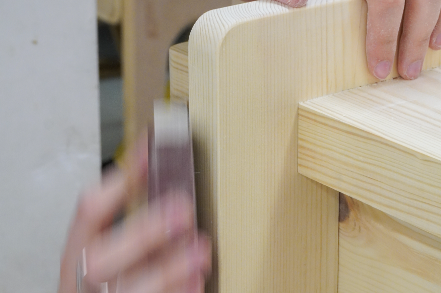 A hand is sanding an item of furniture made of pine.