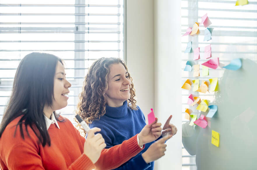 Two people are collaborating in a bright office space, placing colorful sticky notes on a glass wall as part of a brainstorming or planning session.