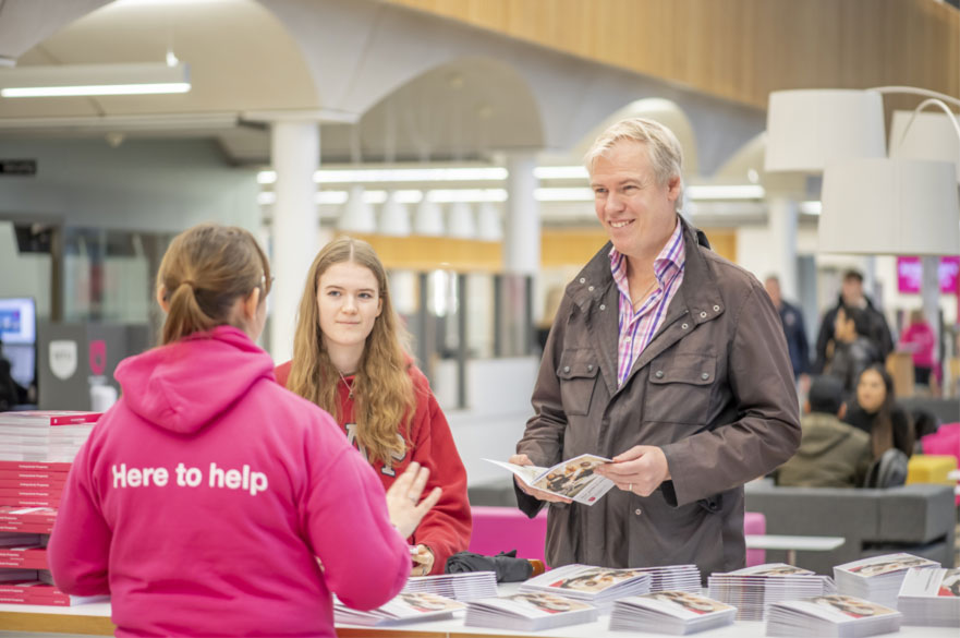 A dad and his daughter talking to a member of staff wearing a pink 'Here to help' hoodie at an open day.