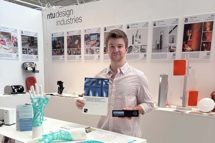 A person holding two awards, stood at a table in front of his work at an exhibition.