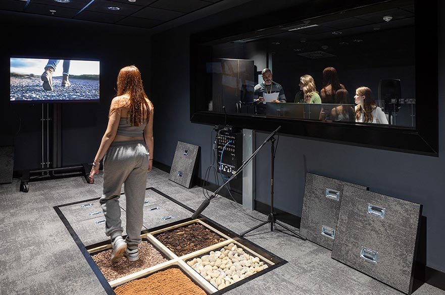 A student walks across different textured Foley surfaces, such as gravel, wood chips, and stones, while being recorded with a microphone, as a group watches through a control-room window in a professional sound studio.
