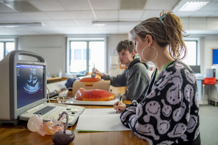 Students operating an ultrasound machine on a training model while the other takes notes in a classroom