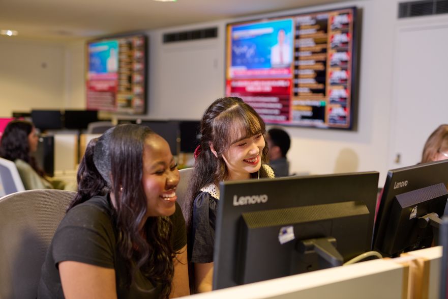 Two students smiling as they look at a computer in the business lab, with stocks and shares showing on the big screen behind them.