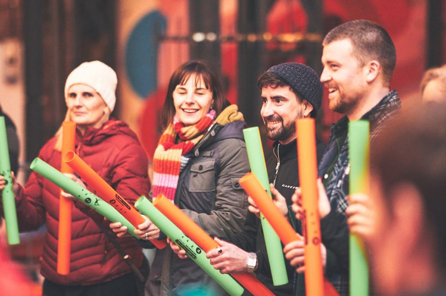 Four people stand in a row. They are all holding two boomwhackers, one in each hand. They are all smiling.