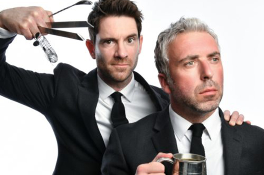 Two men in suits, one holding a drink and the other holding up some cocktail making equipment in his hand.