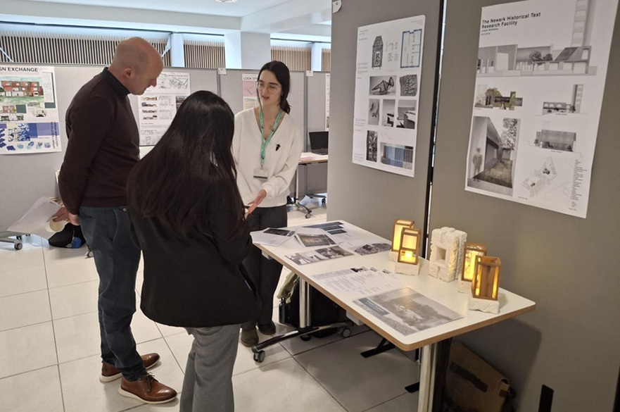 A person showing her work portfolio to other people at an exhibition of student architecture projects.