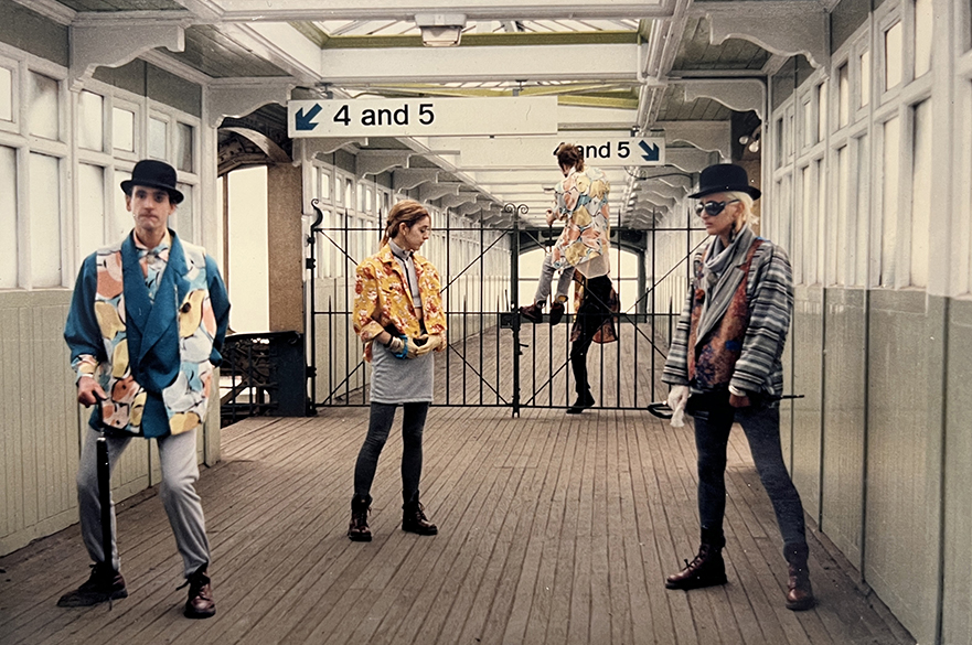 Two men wearing 80s-style loud jackets and bowler hats, with umbrellas, and a women posing on a covered railway station footbridge, whilst two more people are climbing an iron gate in the background.