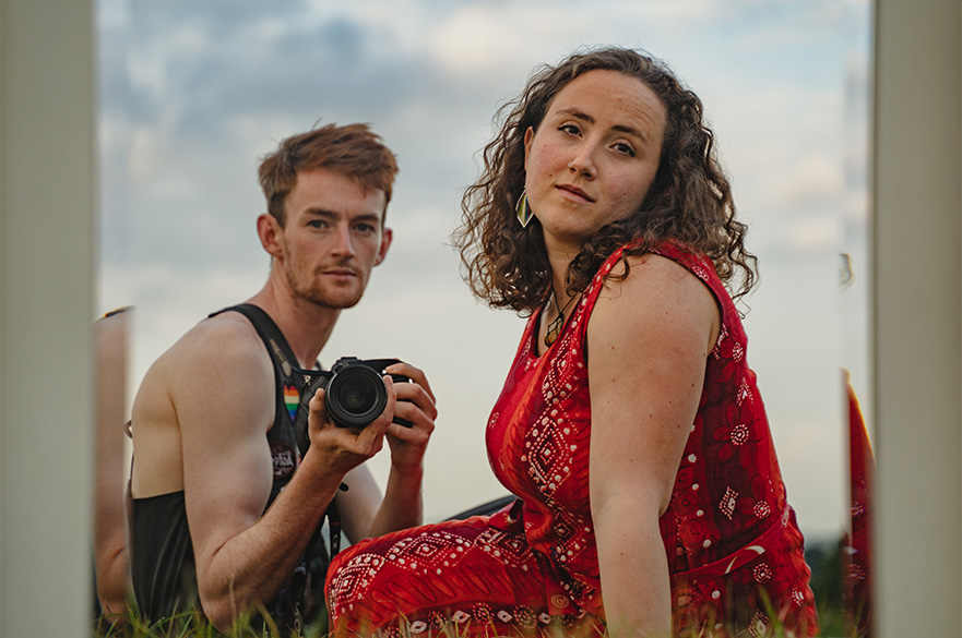 A man holding a camera and a woman in a red dress sat on a field.