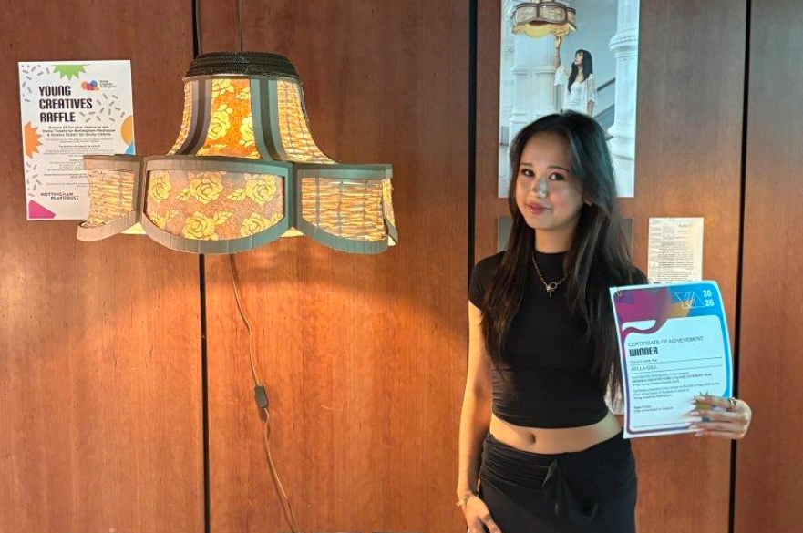A student posing for a photograph, holding an award certificate, standing next to the lampshade they have designed.
