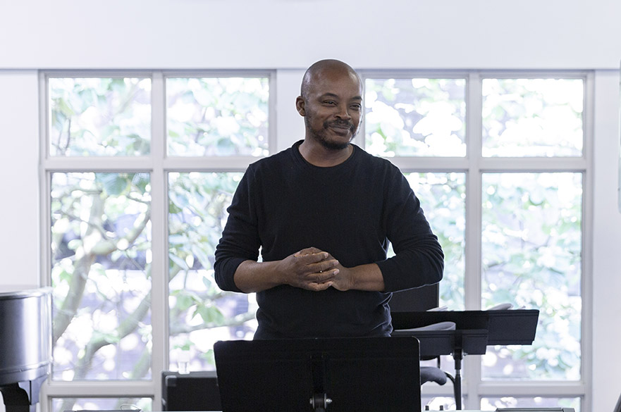 A man who is smiling, standing in front of a window clasping his hands.