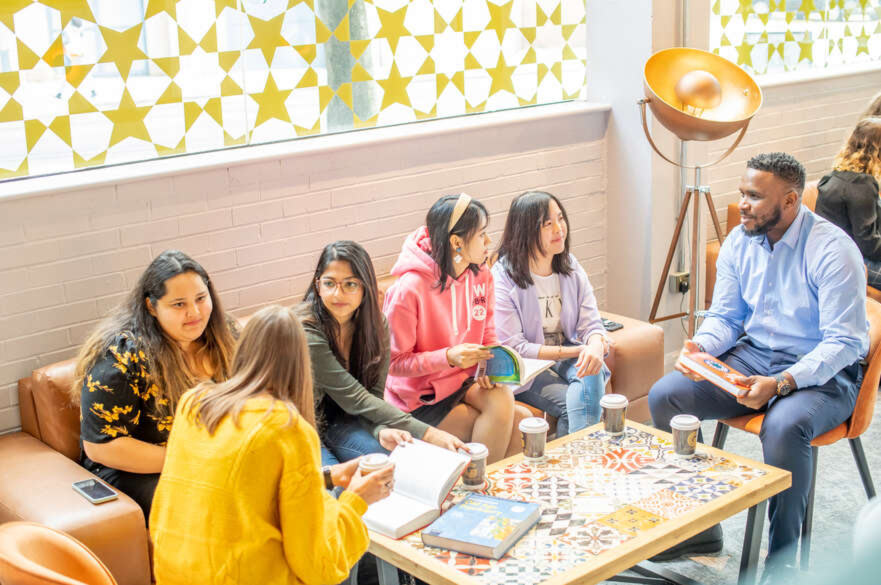 A diverse group of six students gathered around a table in a brightly lit room, chatting and drinking coffee.